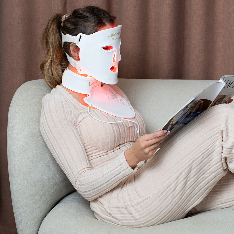 Woman using a LED face and neck mask while reading on a couch.