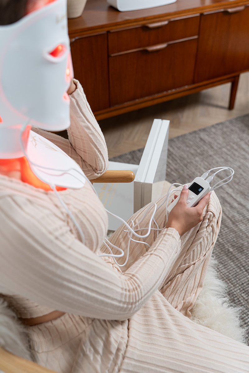 Person sitting in a chair using LED Face mask with visible remote control in a home setting