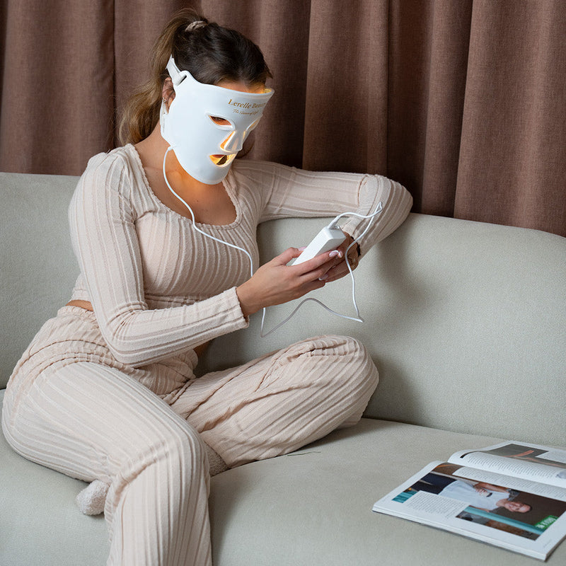 Person wearing led face mask and using a device on a couch with a magazine beside them, against a brown curtain background.